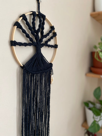 A macrame tree of life wall hanging made on a circular wooden hoop with black knotted yarn, hanging against a beige painted wall. 