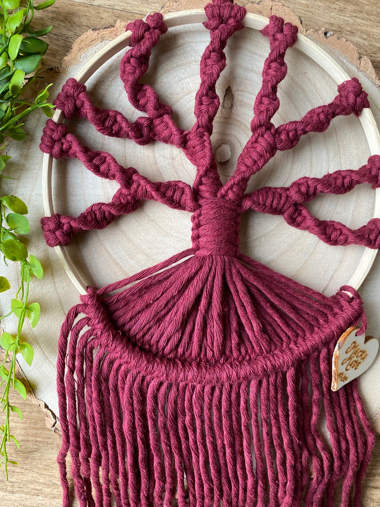 A macrame tree of life wall hanging made on a circular wooden hoop with deep red coloured knotted yarn, laying on a wooden board with some green plant vines. 