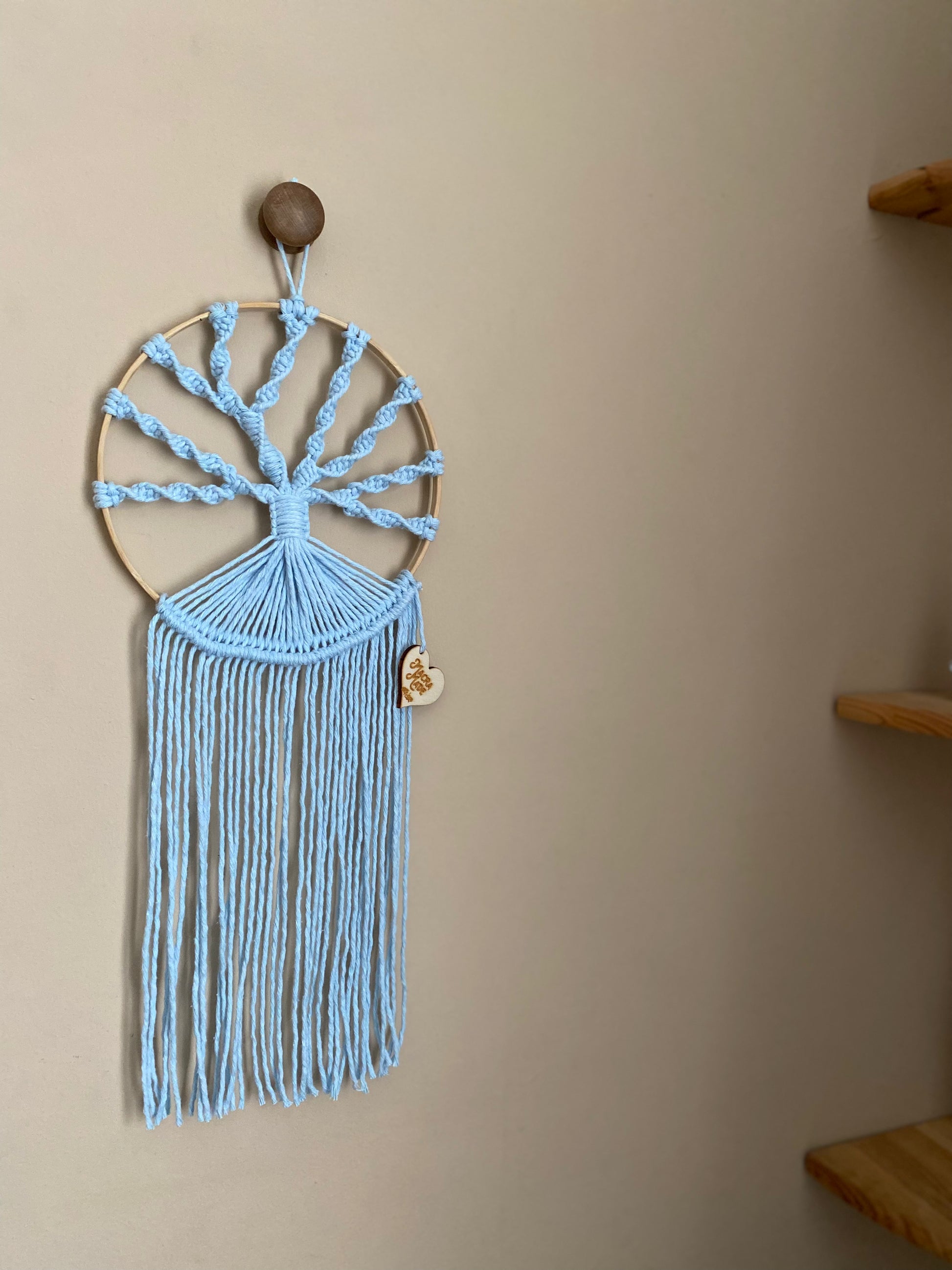A macrame tree of life wall hanging made on a circular wooden hoop with light blue knotted yarn, hanging against a beige painted wall. 