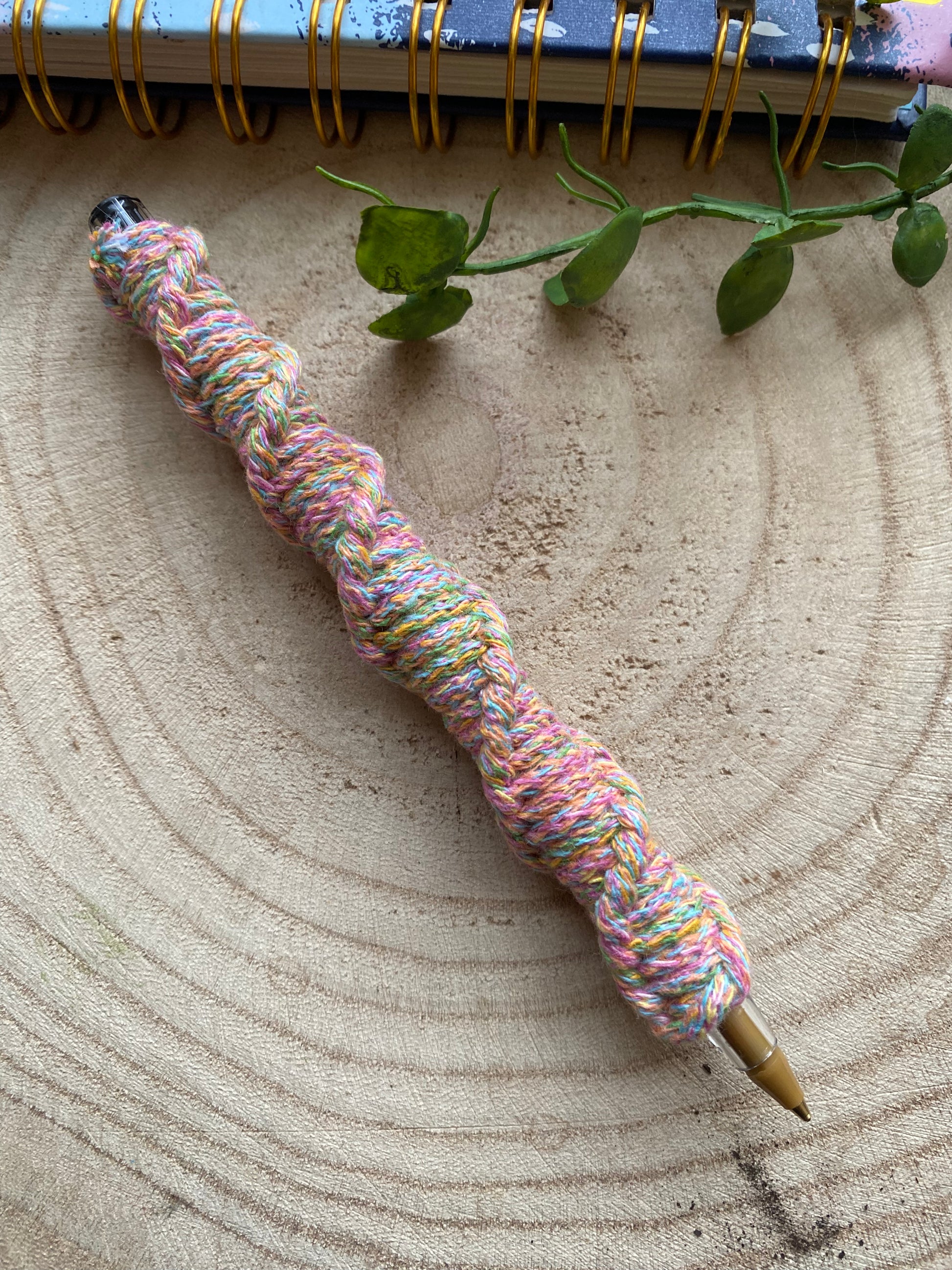 A ballpoint pen wrapped in rainbow coloured yarn with a twisted pattern, laying on a wooden surface. 