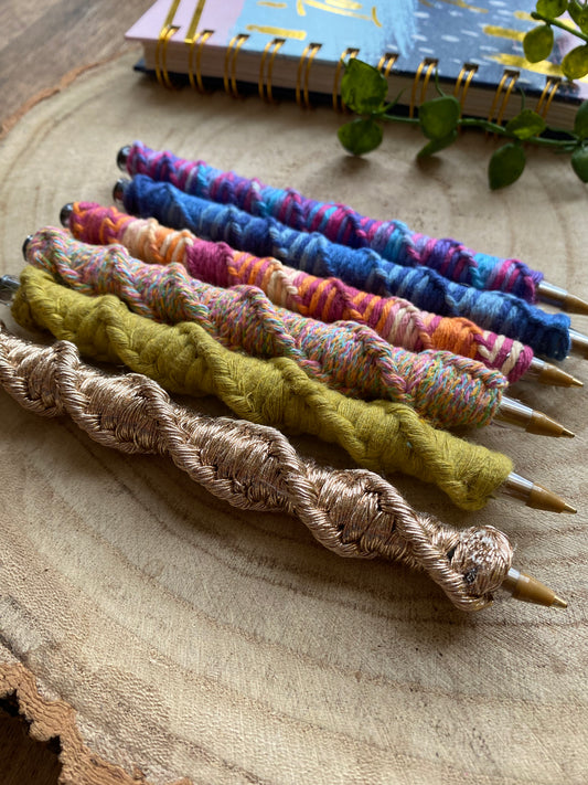 6 ballpoint pens each wrapped in a different colour of yarn with a twisted pattern, laying next to each other on a wooden surface. 