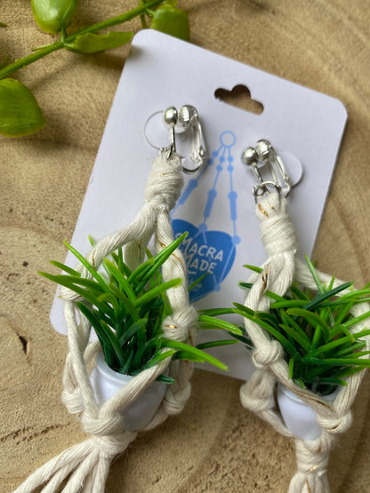 A pair of mini macrame plant hanger earrings made with natural yarn and small artificial plants, with silver clip on hooks, on a white backing card, on a wooden surface. 