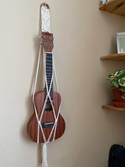 A dark brown ukulele sits inside of a handmade macrame wall hanging ukulele hanger, which is made with natural recycled cotton, and a repurposed wooden hanging ring. 