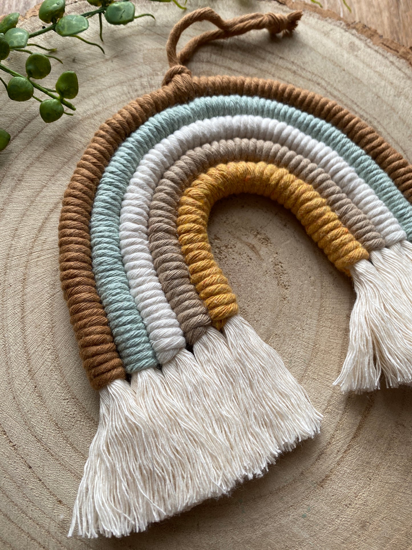 A handmade macrame rainbow, made with coloured yarns in shades of brown, green, white, and mustard yellow, displayed on a wooden surface. 