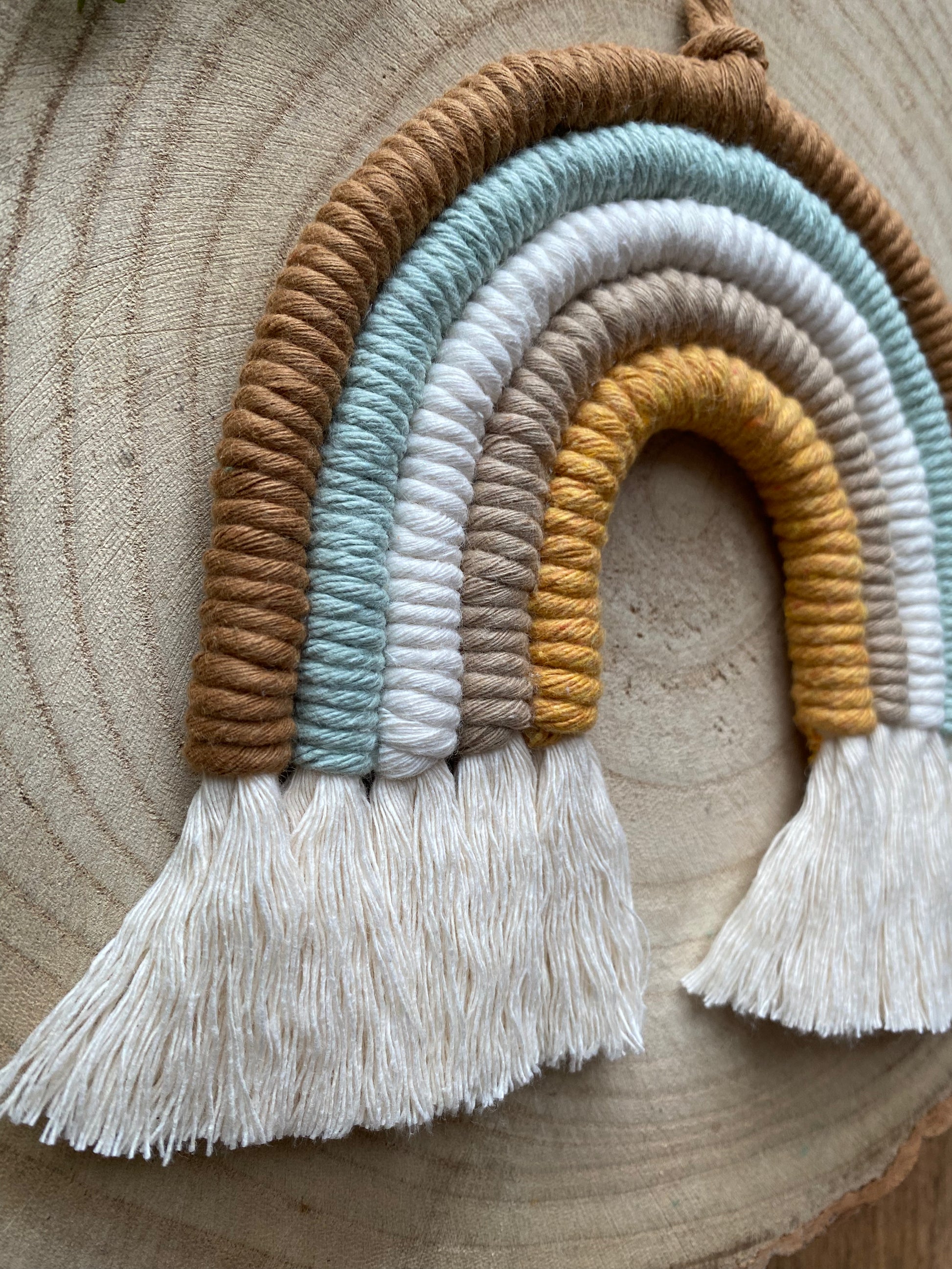 A close up side angled view of a handmade Neutral Rainbow decoration by Macra-Made-With-Love made with coloured yarns in shades of brown, green, white, and mustard yellow, displayed on a wooden surface. 