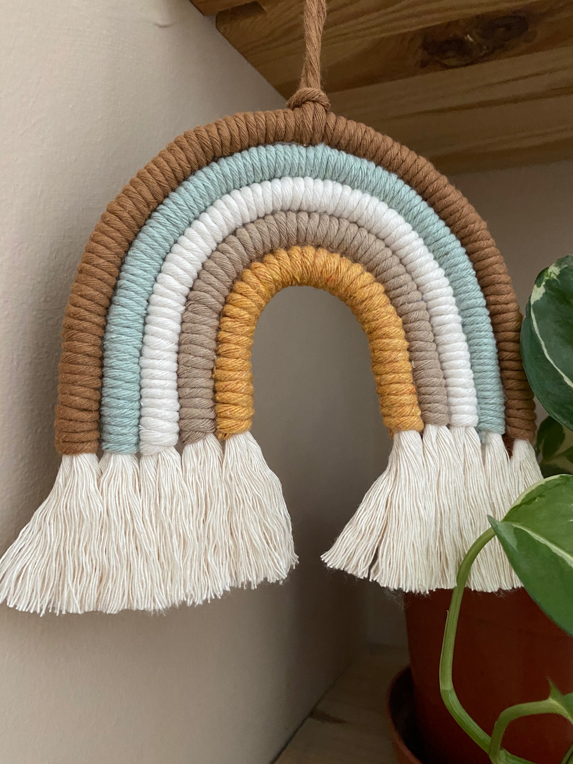 A handmade Neutral Rainbow decoration by Macra-Made-With-Love made with coloured yarns in shades of brown, green, white, and mustard yellow, displayed hanging under a wooden shelf, next to a small variegated houseplant. 