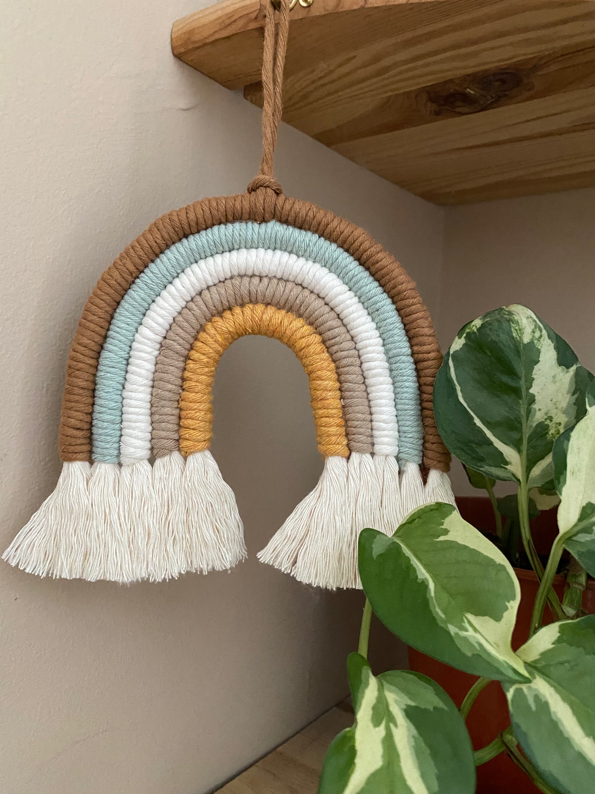 A handmade Neutral Rainbow decoration by Macra-Made-With-Love made with coloured yarns in shades of brown, green, white, and mustard yellow, displayed hanging from a wooden shelf beside a small houseplant. 
