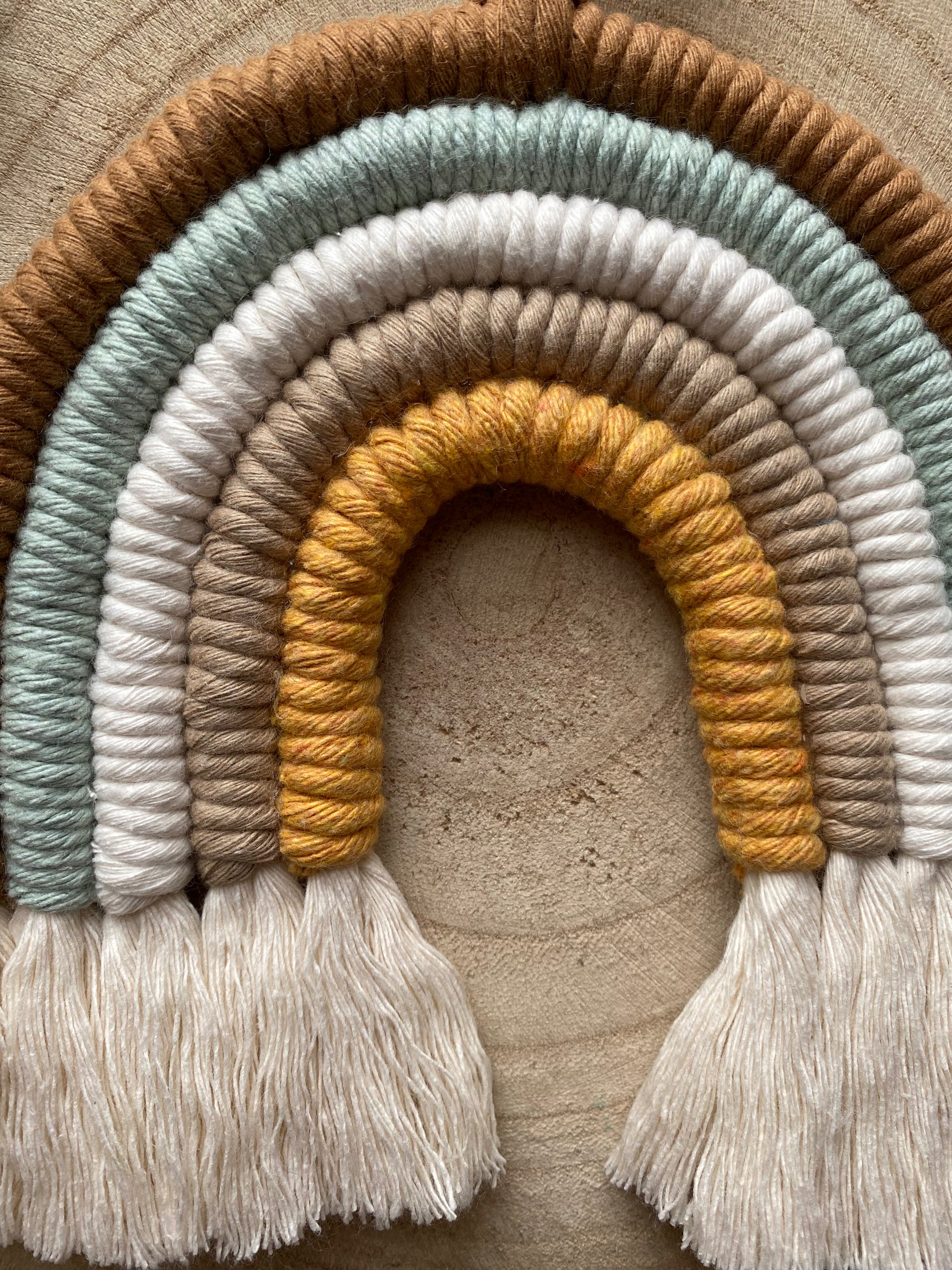 A close up of a handmade Neutral Rainbow decoration by Macra-Made-With-Love made with coloured yarns in shades of brown, green, white, and mustard yellow, displayed on a wooden surface. 