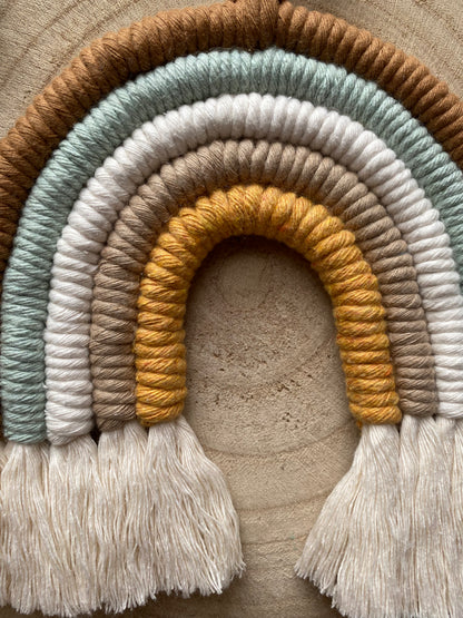 A close up of a handmade Neutral Rainbow decoration by Macra-Made-With-Love made with coloured yarns in shades of brown, green, white, and mustard yellow, displayed on a wooden surface. 