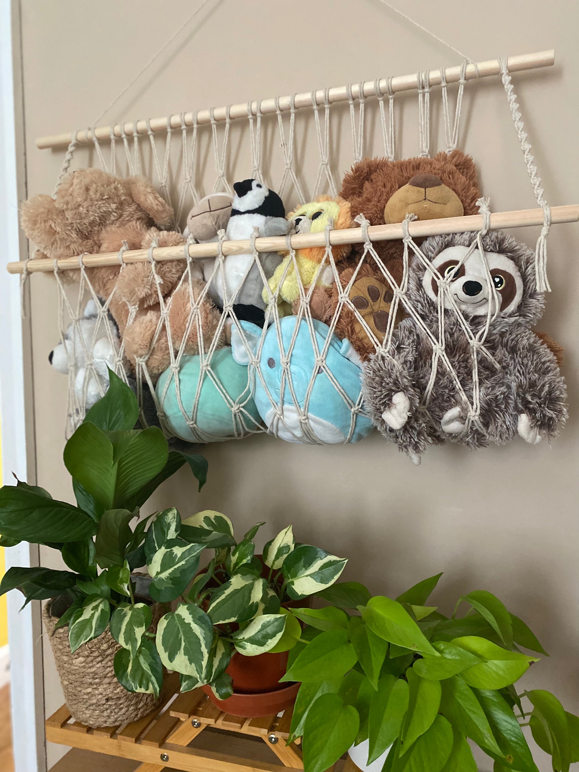 A macrame toy hammock made with natural yarn and wooden dowels, hanging against a beige wall, filled with soft toys and teddies. There are multiple lush green houseplants just below. 