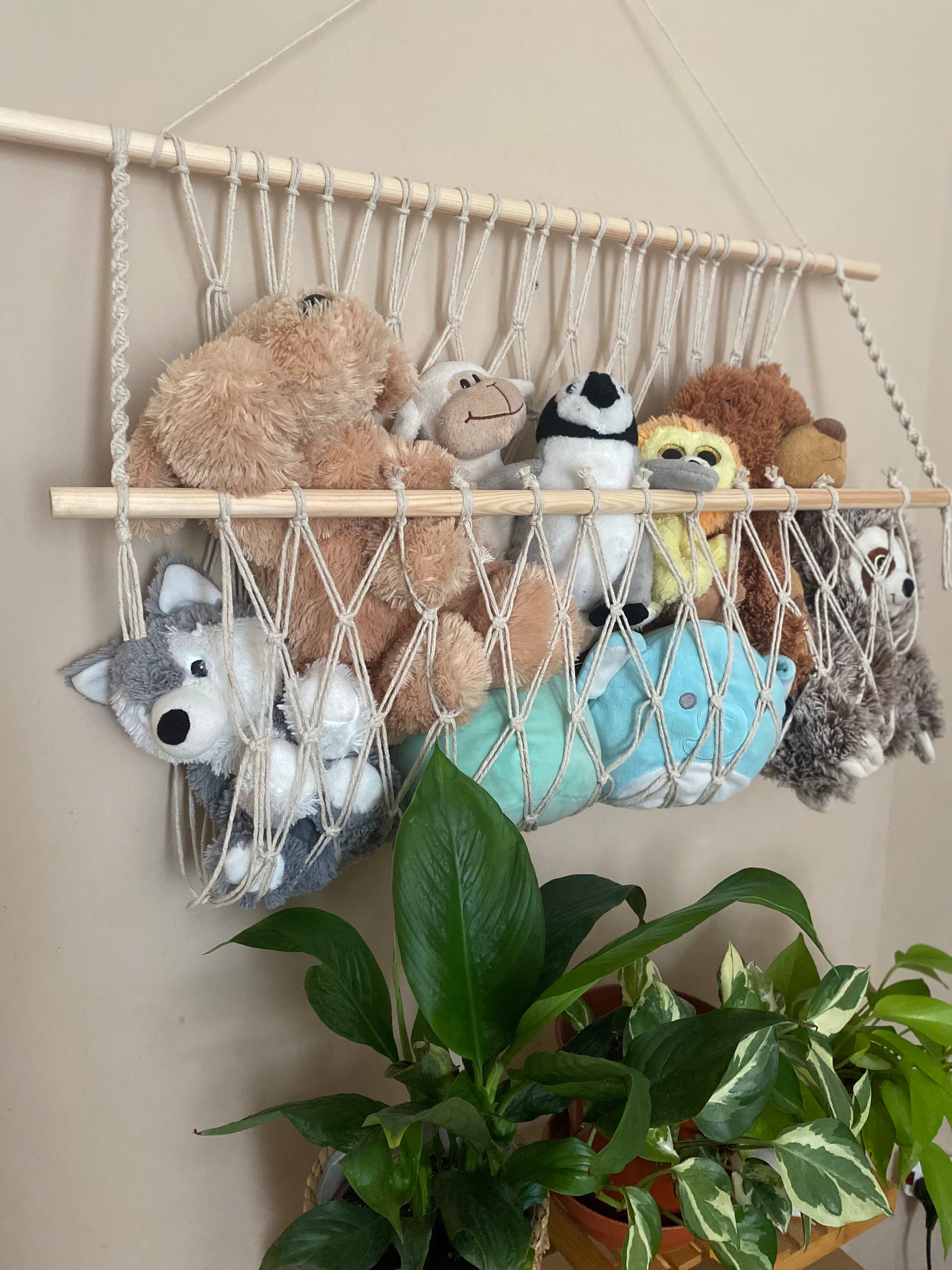 A macrame toy hammock made with natural yarn and wooden dowels, hanging against a beige wall, filled with soft toys and teddies. There are multiple lush green houseplants just below. 