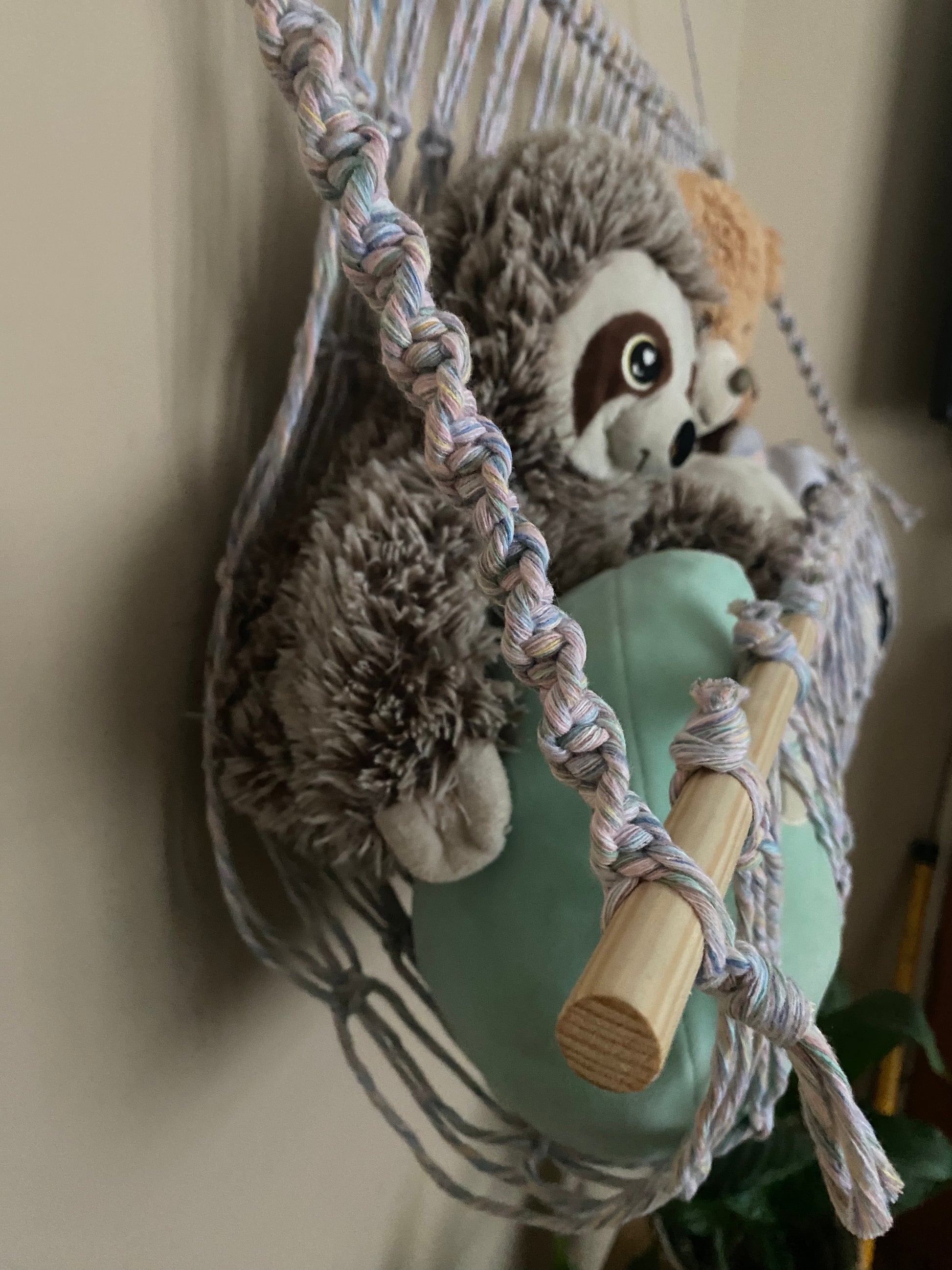 A close up side view of a macrame toy hammock made with pastel coloured yarn and wooden dowels, hanging against a beige wall, filled with soft toys and teddies. 