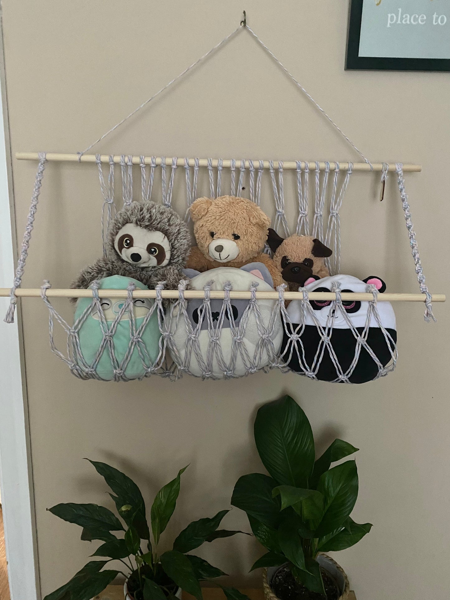 A macrame toy hammock made with pastel yarn and wooden dowels, hanging against a beige wall, filled with soft toys and teddies. There are multiple lush green houseplants just below. 