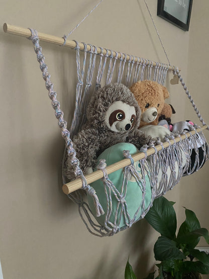 A close up view of a macrame toy hammock made with pastel yarn and wooden dowels, hanging against a beige wall, filled with soft toys and teddies. There are multiple lush green houseplants just below. 