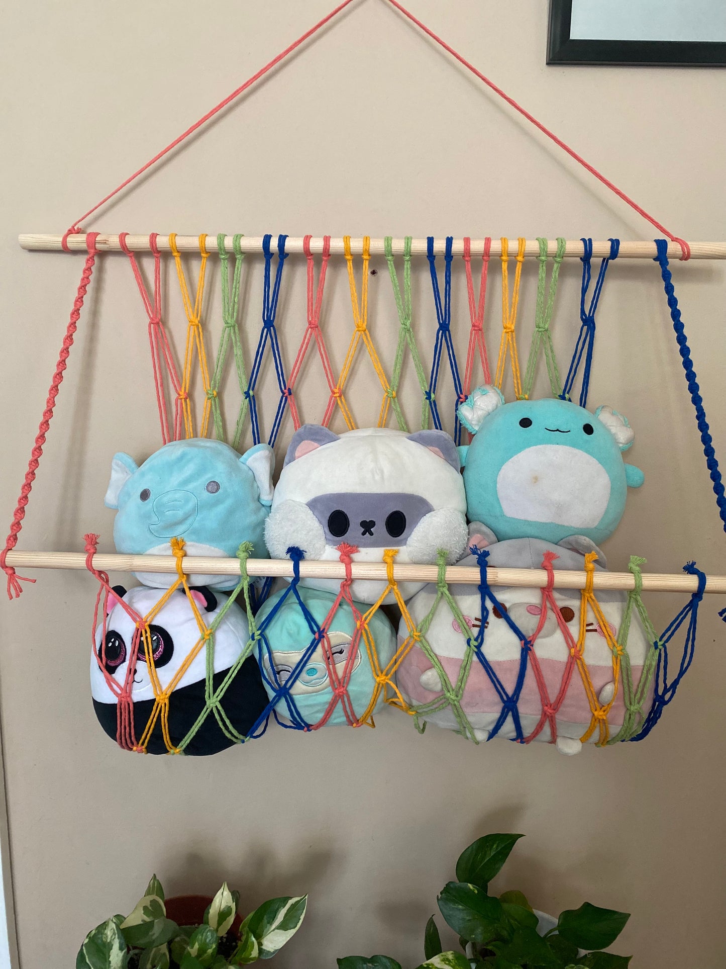 A macrame toy hammock made with colourful yarn and wooden dowels, hanging against a beige wall, filled with soft toys and teddies. There are multiple lush green houseplants just below. 