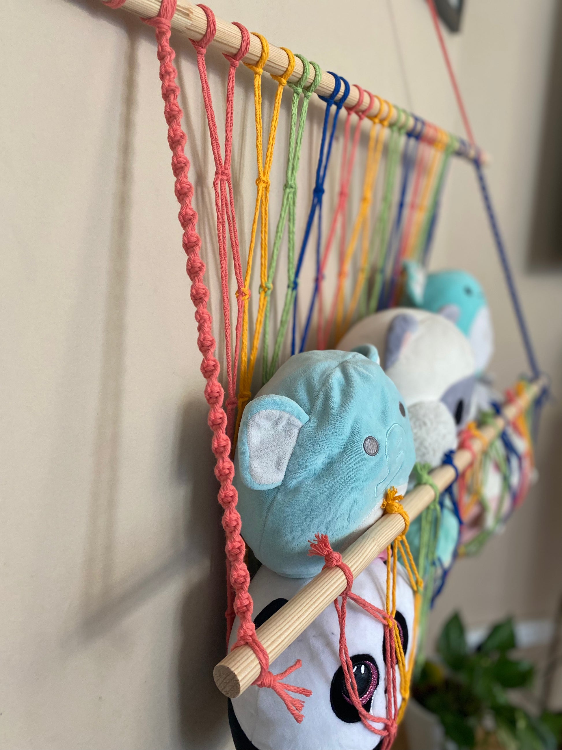 A close up side view of a macrame toy hammock made with colourful yarn and wooden dowels, hanging against a beige wall, filled with soft toys and teddies. 
