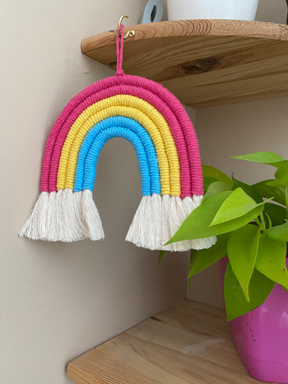 A macrame rainbow made with 6 arches of pink, yellow and blue yarn to represent the colours of the pansexual pride flag, with combed out tassels at the bottom, hanging on a wooden corner shelf next to a bright green houseplant. 