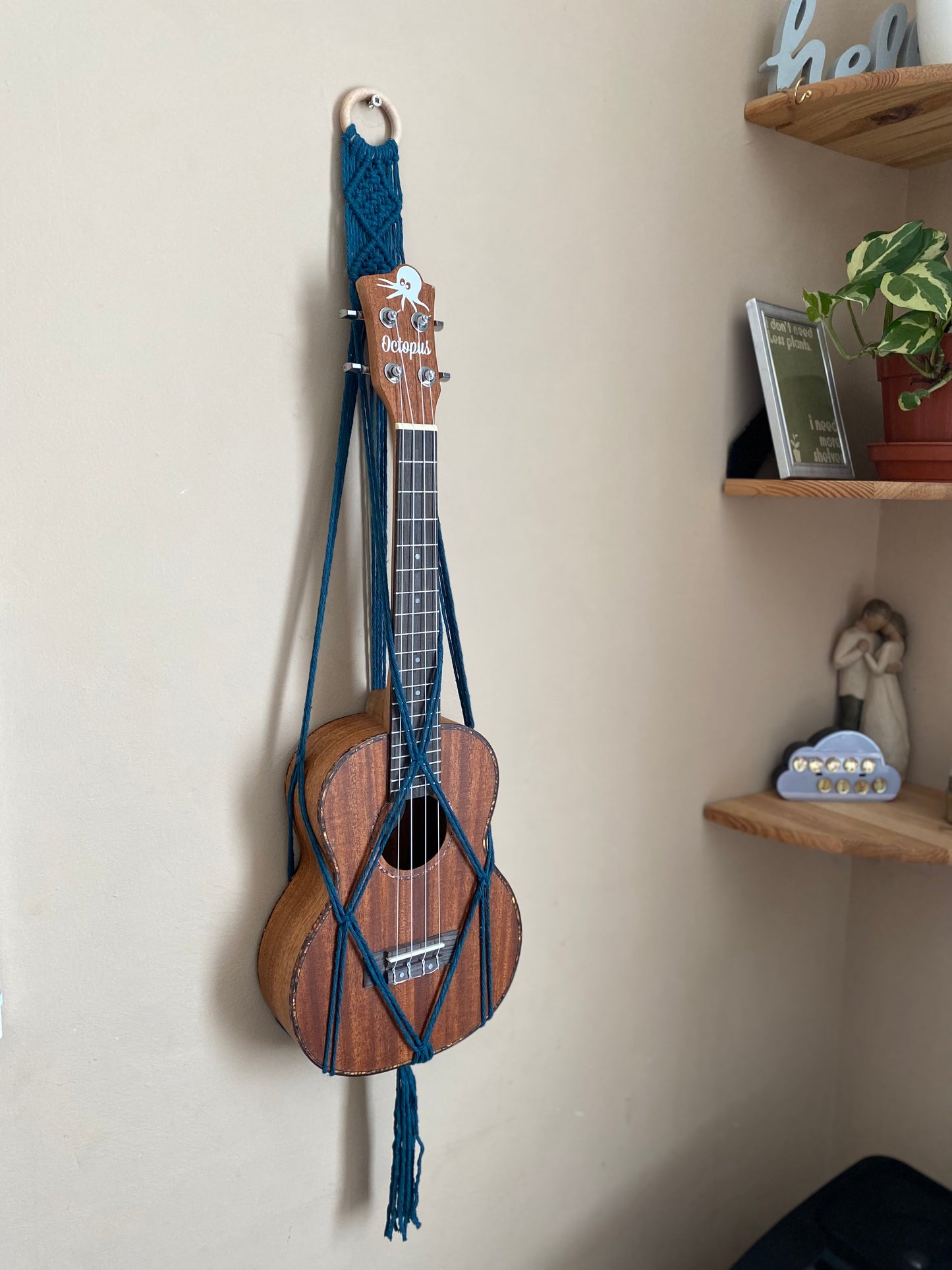 A dark brown ukulele sits inside of a handmade macrame wall hanging ukulele hanger, which is made with petrol blue recycled cotton, and a repurposed wooden hanging ring. A set of corner shelves are to the right of the image. 
