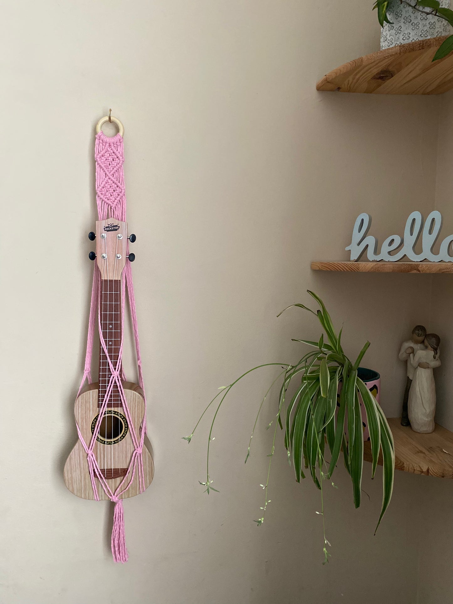 A light brown ukulele sits inside of a handmade macrame wall hanging ukulele hanger, which is made with light pink recycled cotton, and a repurposed wooden hanging ring. A large spider plant sits on a corner shelf next to the hanger.