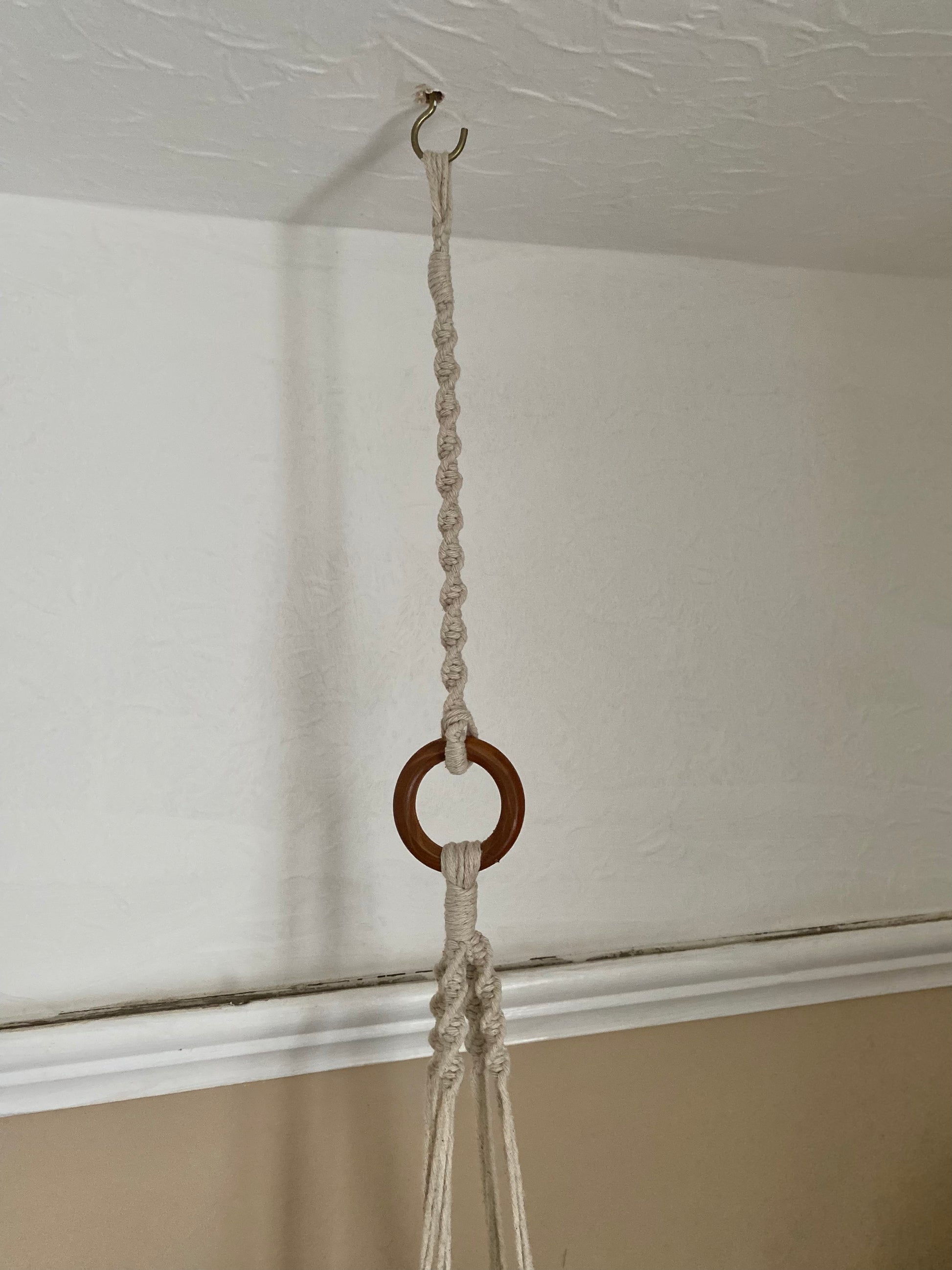 A macrame plant hanger extension made with natural cotton yarn, is attached to the wooden ring of a macrame plant hanger, and suspended from a white ceiling from a metal hook. 