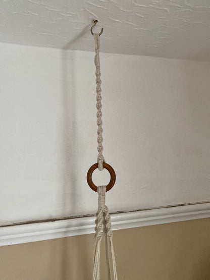 A macrame plant hanger extension made with natural cotton yarn, is attached to the wooden ring of a macrame plant hanger, and suspended from a white ceiling from a metal hook. 