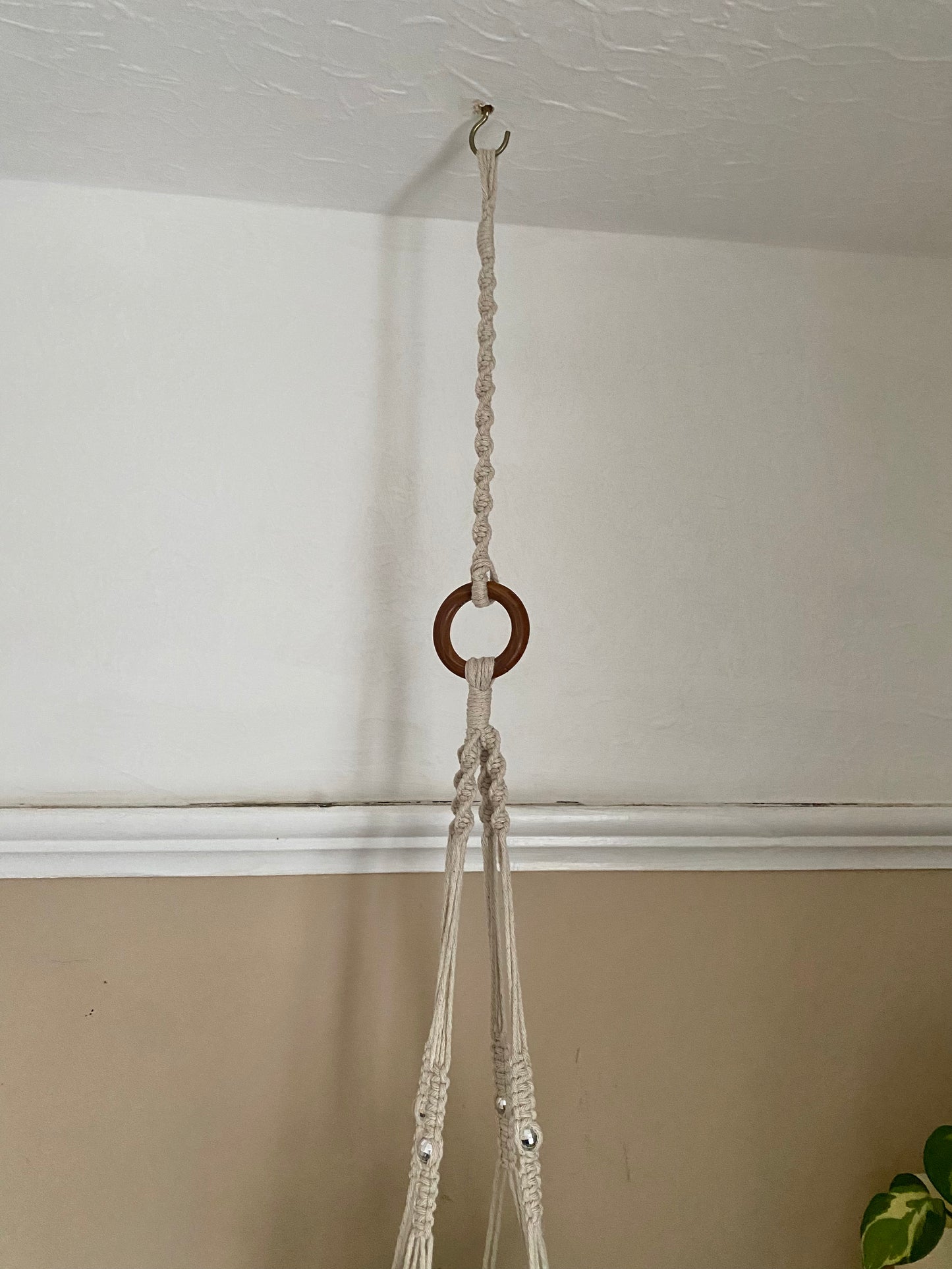 A macrame plant hanger extension made with natural cotton yarn, is attached to the wooden ring of a macrame plant hanger, and suspended from a white ceiling on a metal hook. 