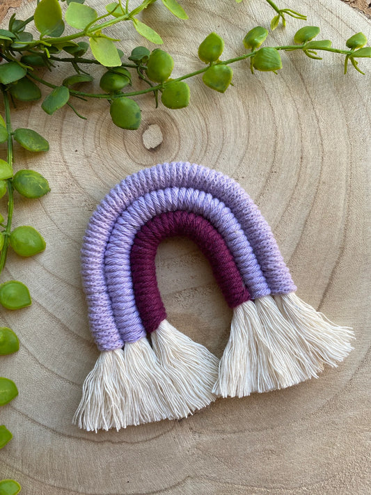 Small handmade macrame rainbow fridge magnet in soft cotton purple cords, on a wooden surface with some green plant vines. 