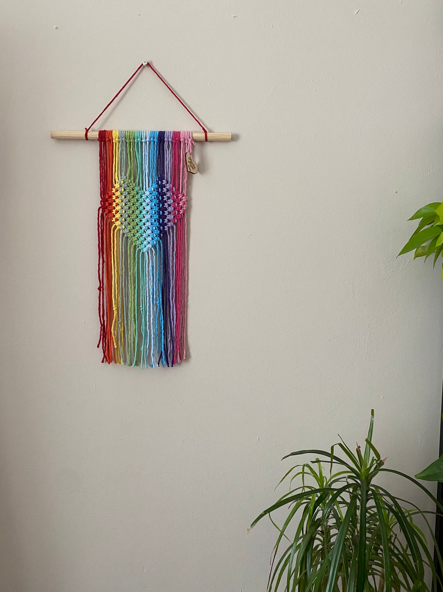 A macrame wall hanging with heart detail, made with rainbow colours of yarn on a wooden dowel, hanging on a beige wall. 