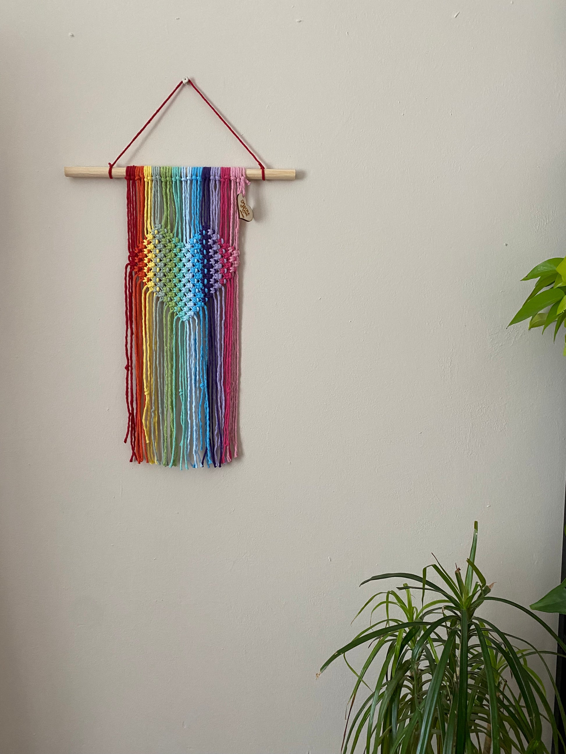 A macrame wall hanging with heart detail, made with rainbow colours of yarn on a wooden dowel, hanging on a beige wall. 