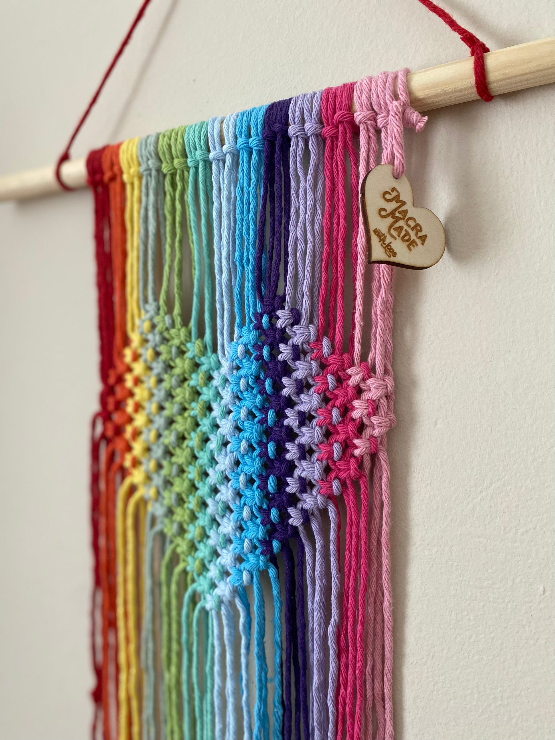 A close up image of a macrame wall hanging with heart detail, made with rainbow colours of yarn on a wooden dowel, hanging on a beige wall. 
