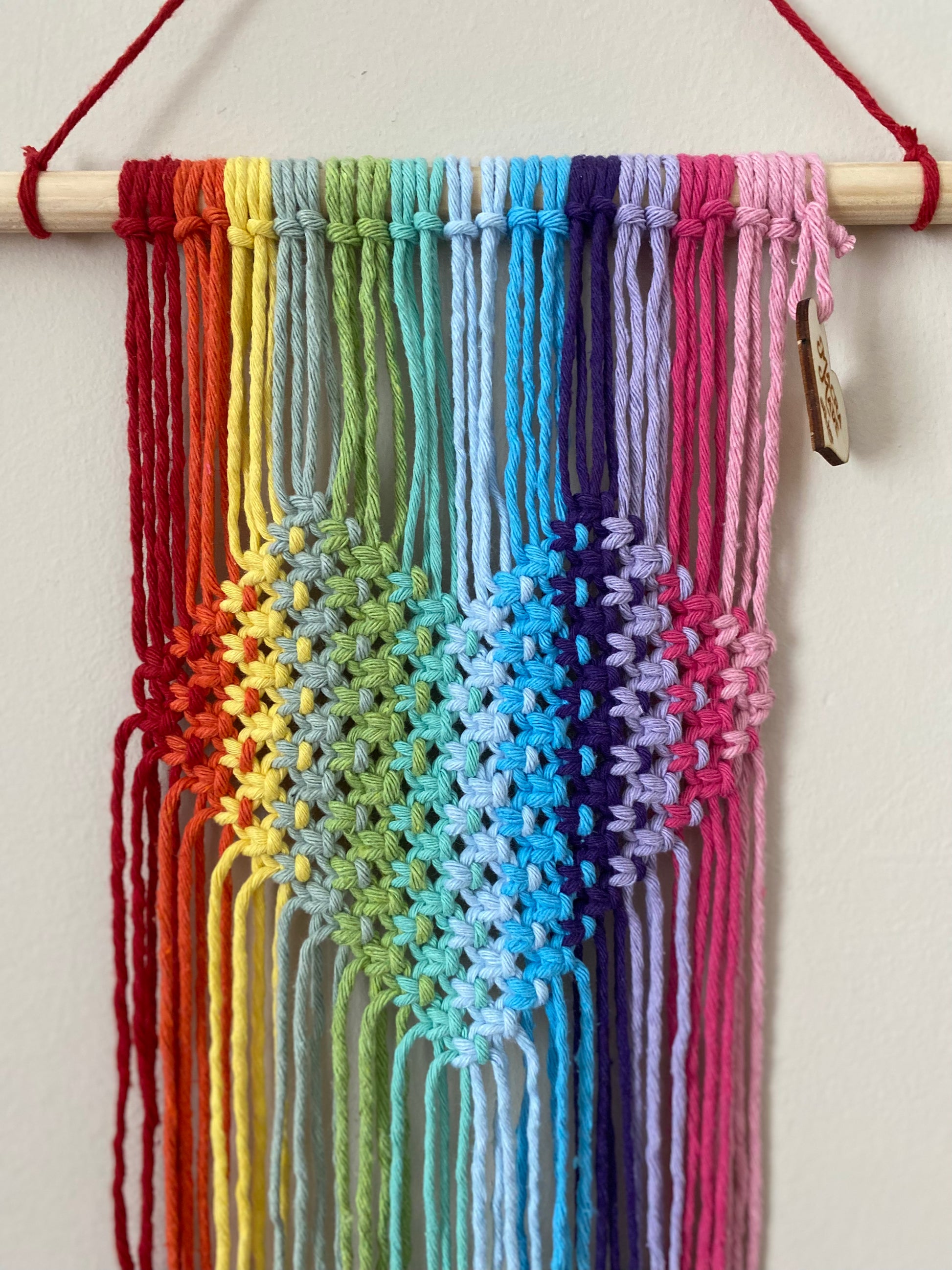 A close up of a macrame wall hanging with heart detail, made with rainbow colours of yarn on a wooden dowel, hanging on a beige wall. 