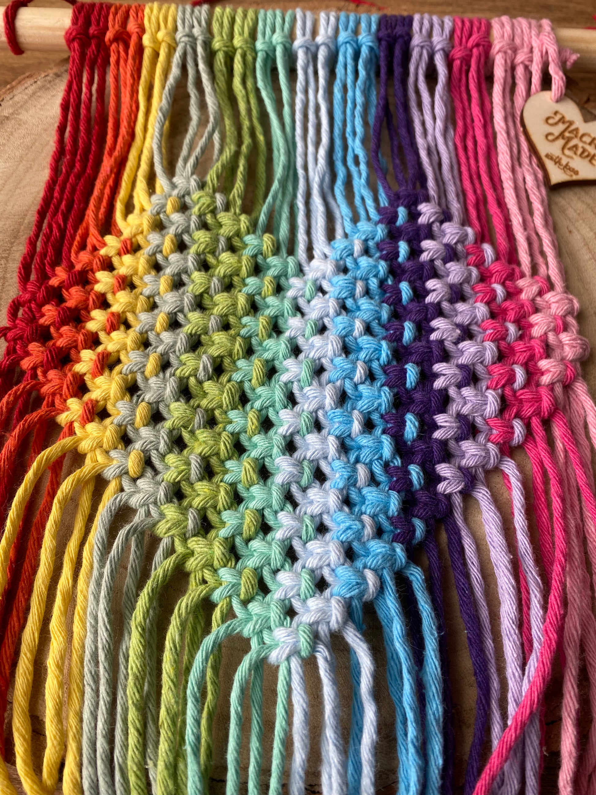 A close up of a macrame wall hanging with heart detail, made with rainbow colours of yarn on a wooden dowel, laid out on a wooden surface.