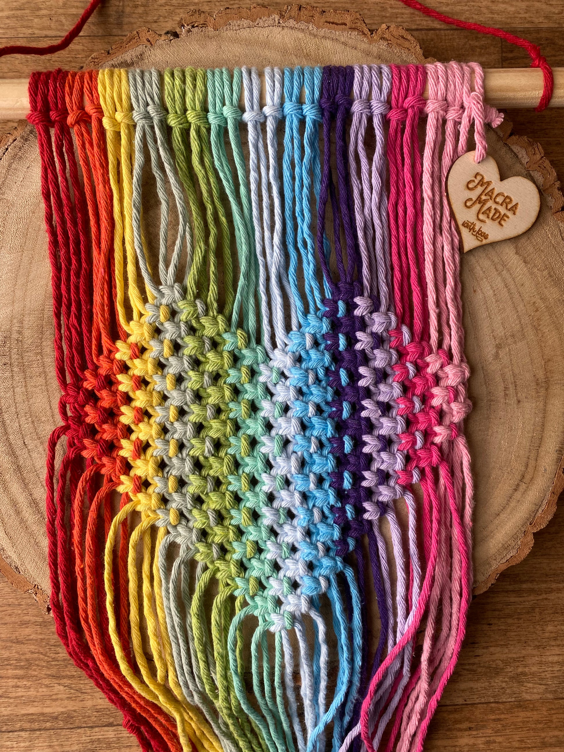 Close up top down view of a macrame wall hanging with heart detail, made with rainbow colours of yarn on a wooden dowel, laid out across a wooden surface. 