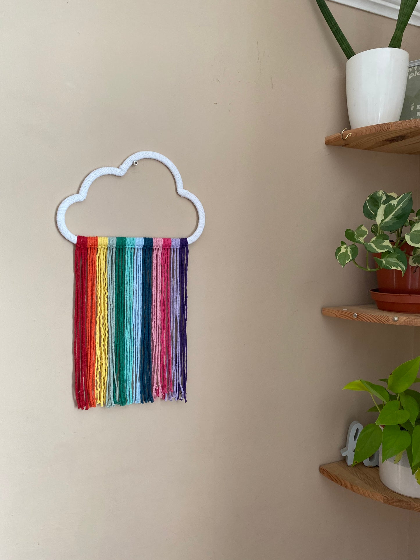 A cloud shaped wall hanging decoration, made with rainbow hanging tassels, displayed against a beige wall. There are corner shelves with houseplants on, in the corner .