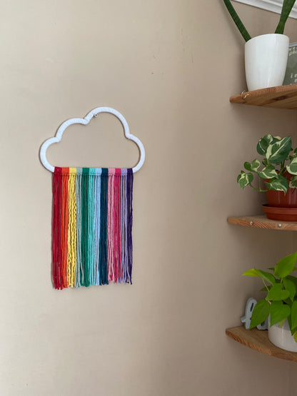 A cloud shaped wall hanging decoration, made with rainbow hanging tassels, displayed against a beige wall. There are corner shelves with houseplants on, in the corner .