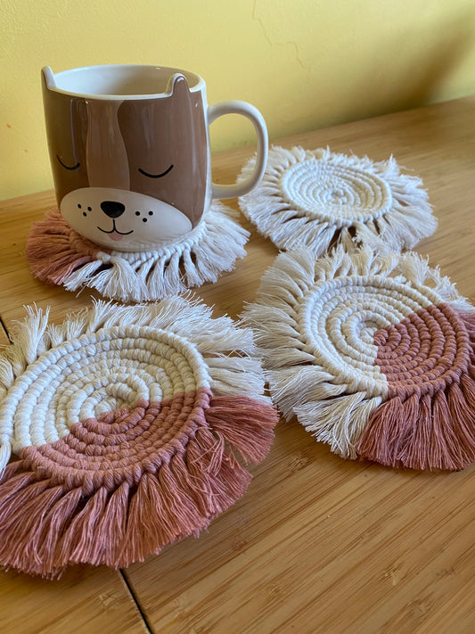 A set of 4 round macrame coasters made with blush pink and natural yarns, with a soft fringed edge, are placed on a wooden table,  with a novelty dog mug placed on top of one of the coasters. 