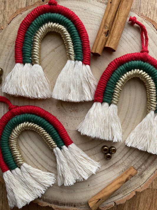 A set of three handmade macrame rainbow Christmas decorations in red, green, and gold yarn with white tassels, displayed on a wooden surface with beads and cinnamon sticks for decoration. 