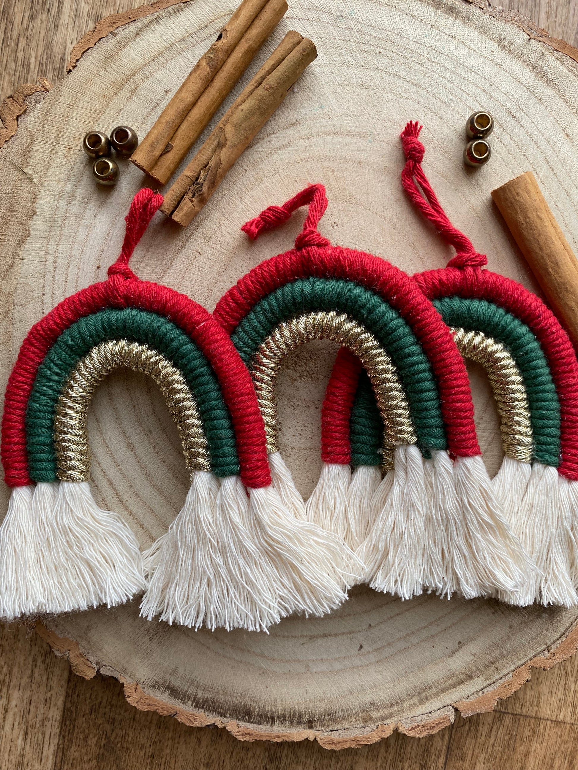 A set of three handmade macrame rainbow Christmas decorations in red, green, and gold yarn with white tassels, displayed next to each other on a wooden surface with beads and cinnamon sticks for decoration. 