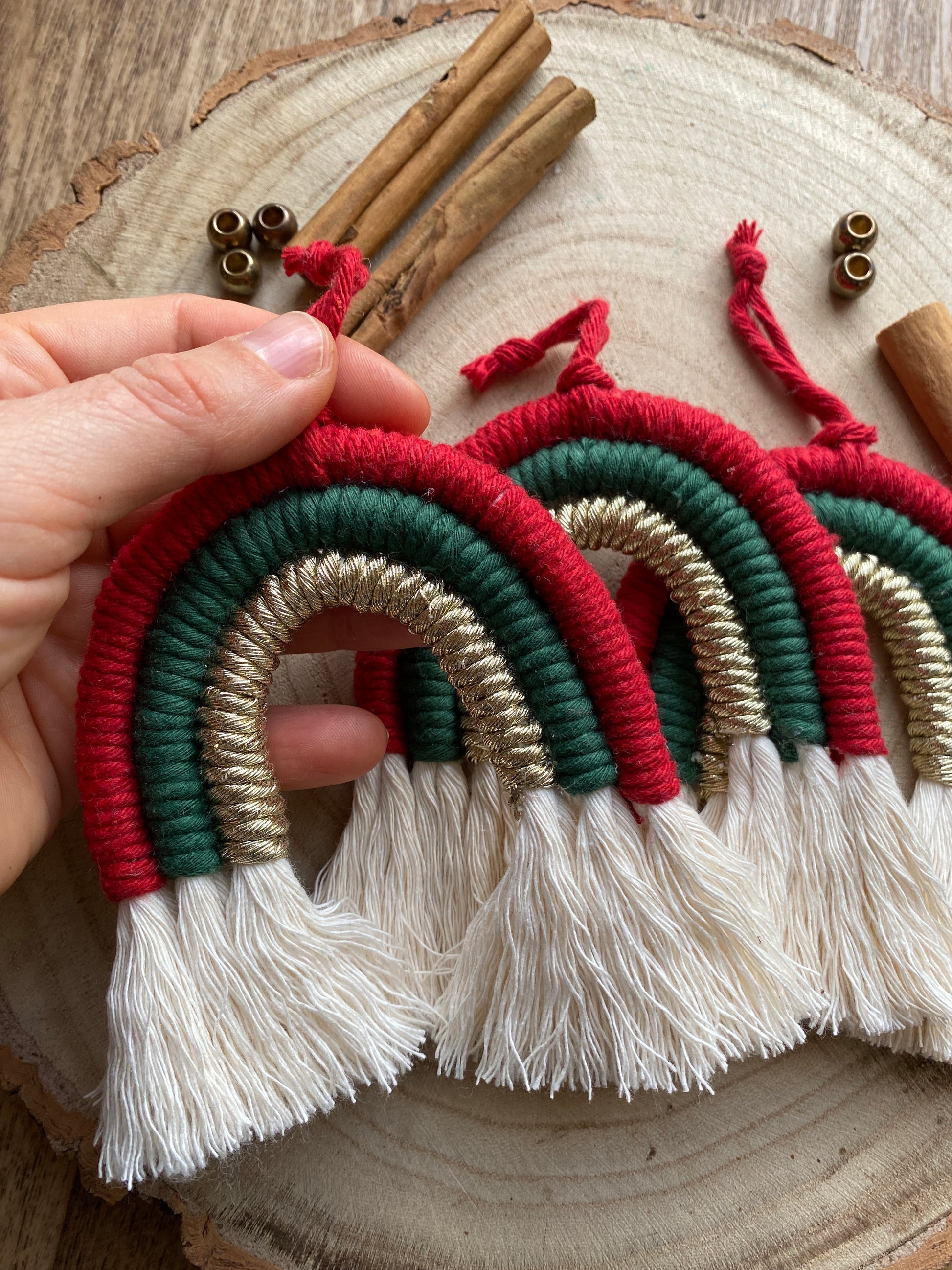 A hand holding one of a set of three handmade macrame rainbow Christmas decorations in red, green, and gold yarn with white tassels, displayed on a wooden surface with beads and cinnamon sticks for decoration. 