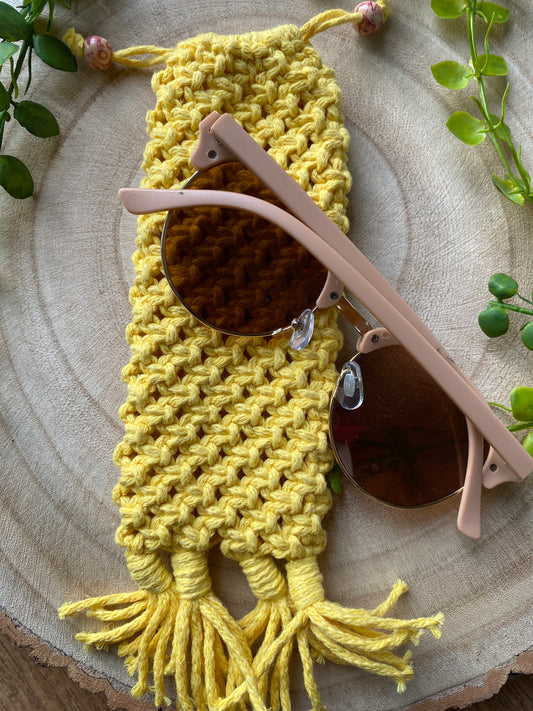 Handmade macrame soft glasses pouch made with yellow cotton yarn. Displayed with a pair of pink sunglasses laid across it, on a round wooden surface. 