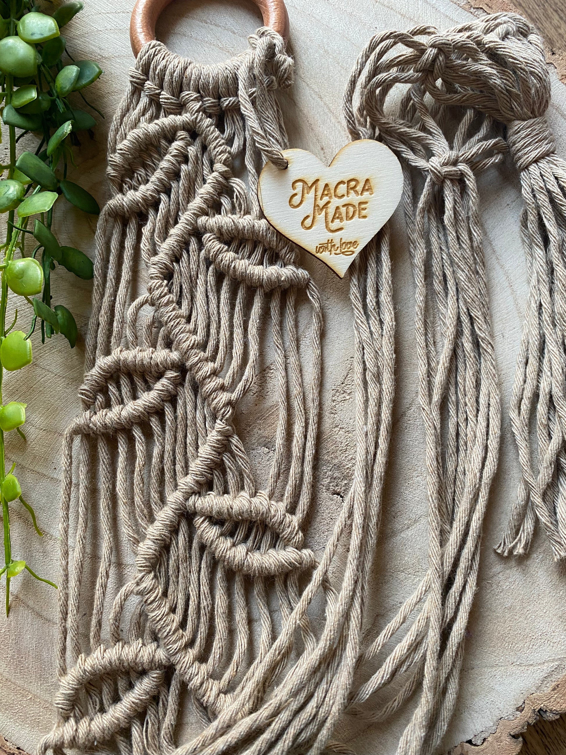 Close up of a neutral coloured macrame plant hanger with falling leaf knotted pattern and a wooden ring, folded neatly on a wooden surface. 
