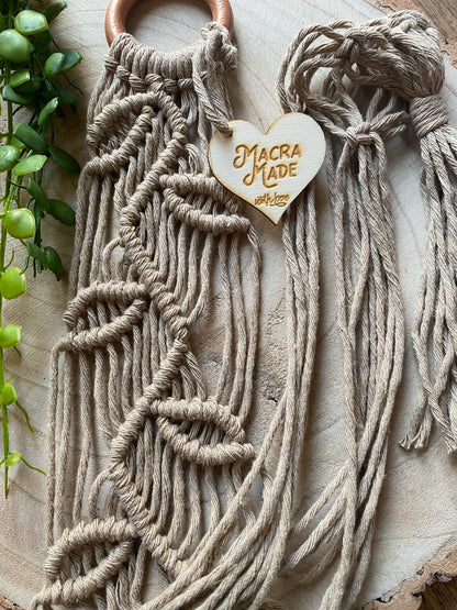 Close up of a neutral coloured macrame plant hanger with falling leaf knotted pattern and a wooden ring, folded neatly on a wooden surface. 