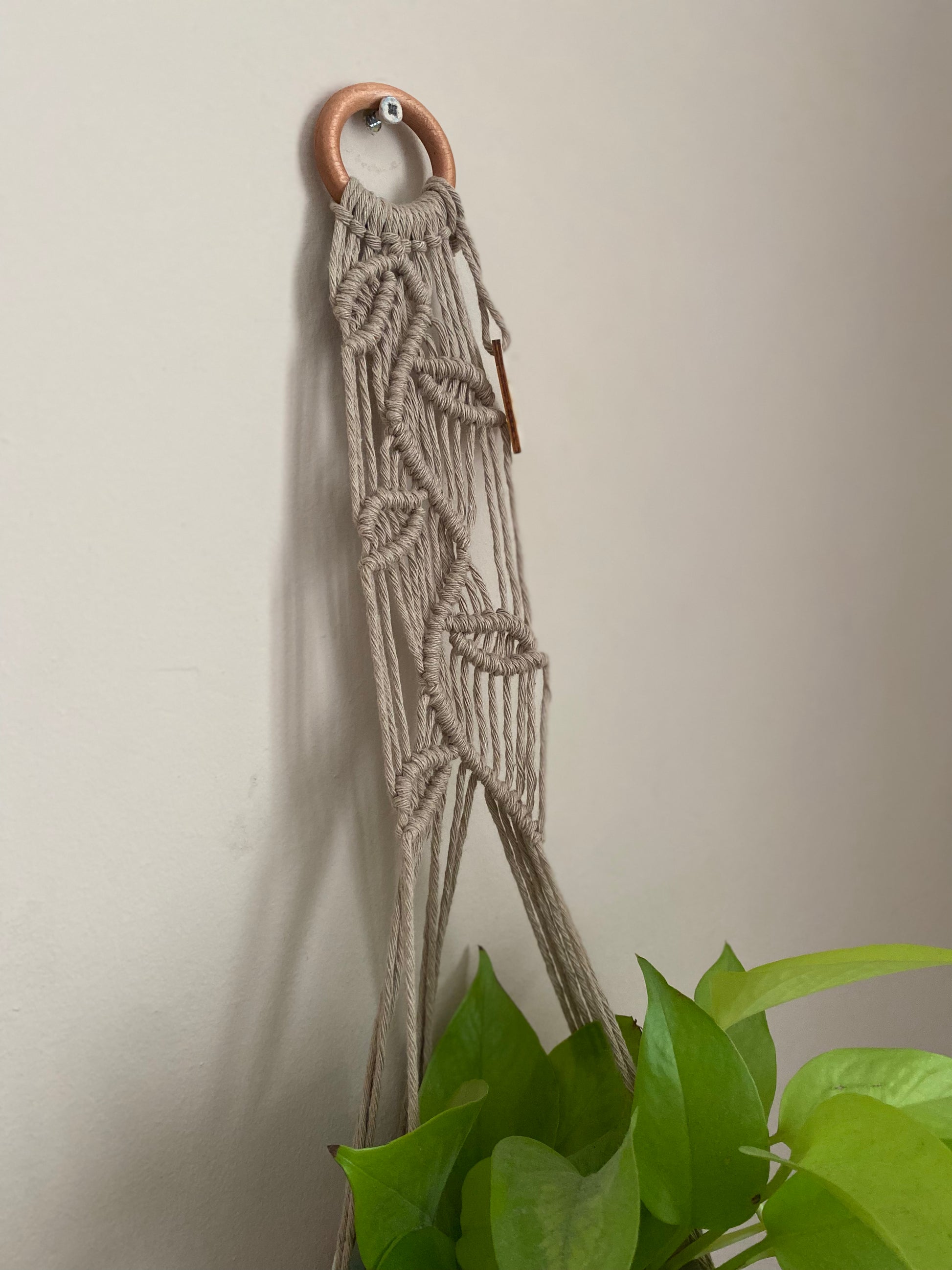 Close up of a neutral coloured macrame plant hanger with falling leaf knotted pattern and a wooden ring hanging against a beige wall. 