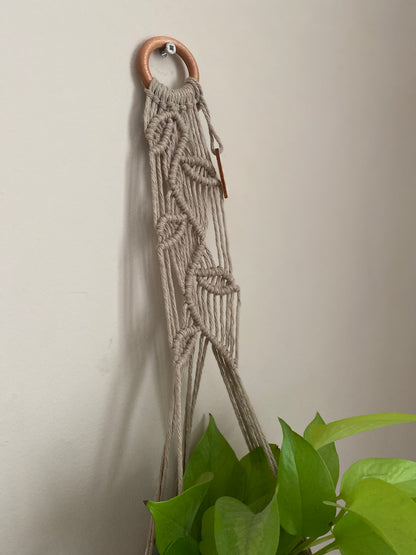Close up of a neutral coloured macrame plant hanger with falling leaf knotted pattern and a wooden ring hanging against a beige wall. 