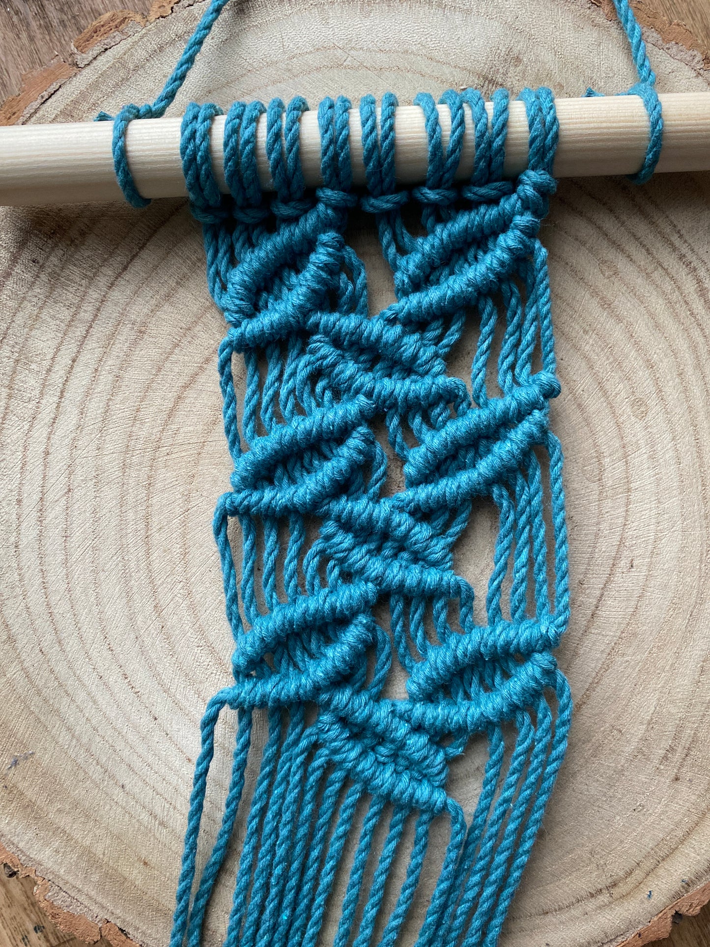 A handmade macrame plant hanger made with teal blue recycled cotton on a wooden dowel, with an intricate falling leaf pattern, laid out on a wooden surface. 