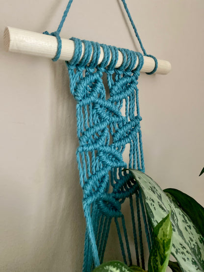 A close up view of a handmade macrame wall hanging plant hanger, made with teal blue recycled cotton on a solid wooden dowel. 
