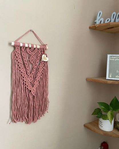 A textured, layered macrame wall hanging made with intricate knot patterns using blush pink yarn on a wooden dowel, hanging against a beige wall next to a set of corner shelves. 
