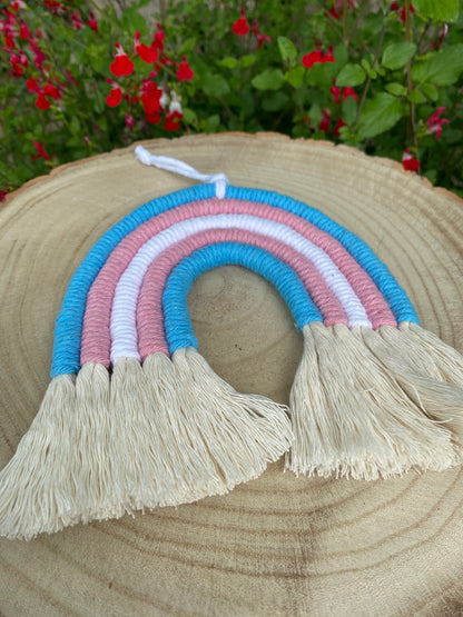 Handmade macrame rainbow made with 5 arches of recycled cotton in blue, pink and white yarns to represent the colours of the trans pride flag, with combed out tassels at the bottom, displayed on a wooden board. 