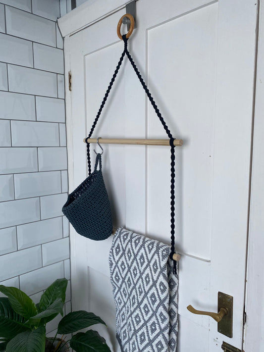 A grey basket and a grey and white patterned towel, hanging on the wooden rods of a two tiered macrame bathroom towel holder, on the back of a white door. 