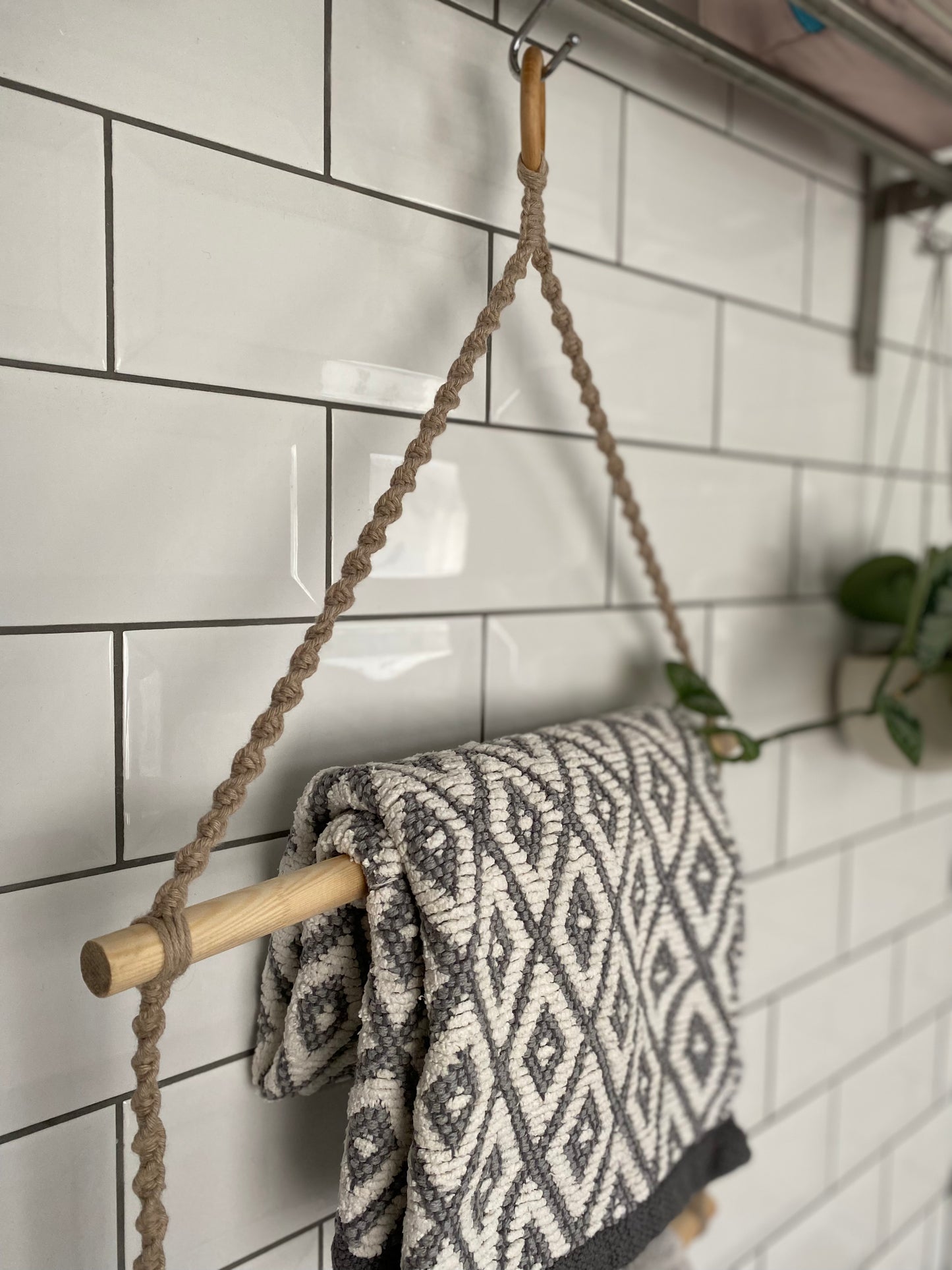 Close up of a bathroom towel holder, made with taupe yarn, wooden dowels and a wooden ring, hanging against a white tiled wall, holding a grey and white patterned towel. 